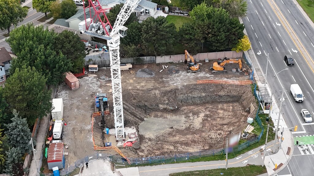 An angled aerial view of the excavated lot, with the crane mast rising from the pit, two excavators along the back fence and site trailers along the side.