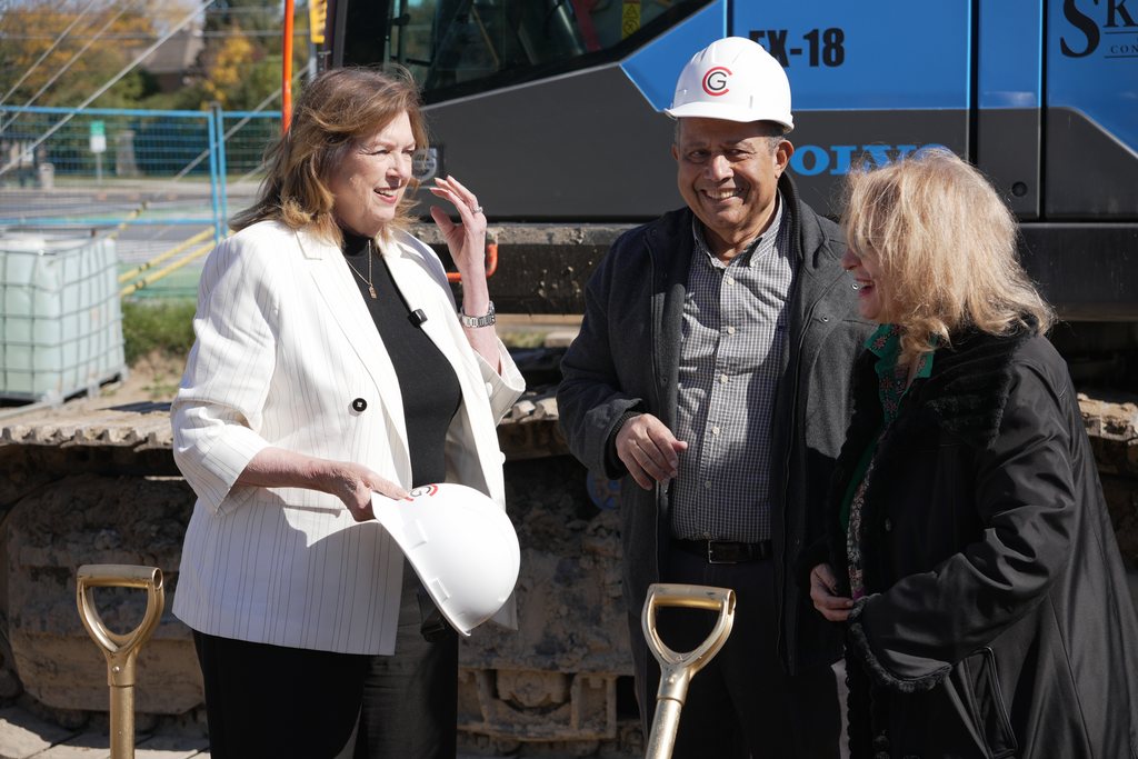 Three guests in hard hats laughing together beside the excavator, gold shovels at their feet.
