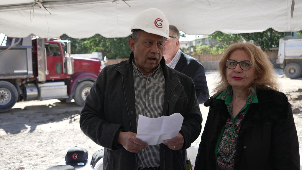 A man in a white hard hat speaks from his notes under the event tent, beside a woman listening, with dump trucks behind.