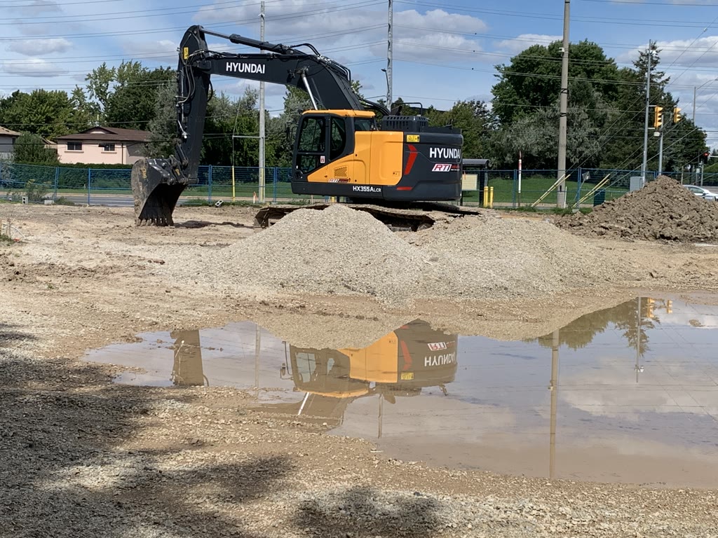 An excavator stands on the cleared corner lot beside piles of gravel and soil, reflected in a puddle, with the street and houses behind.
