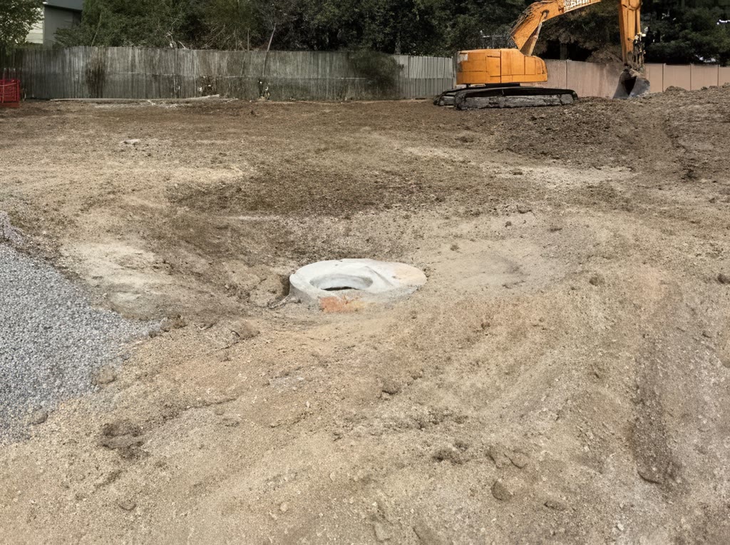 The storm sewer trenches filled back in and levelled, with a manhole cover at grade and an excavator at the far fence.